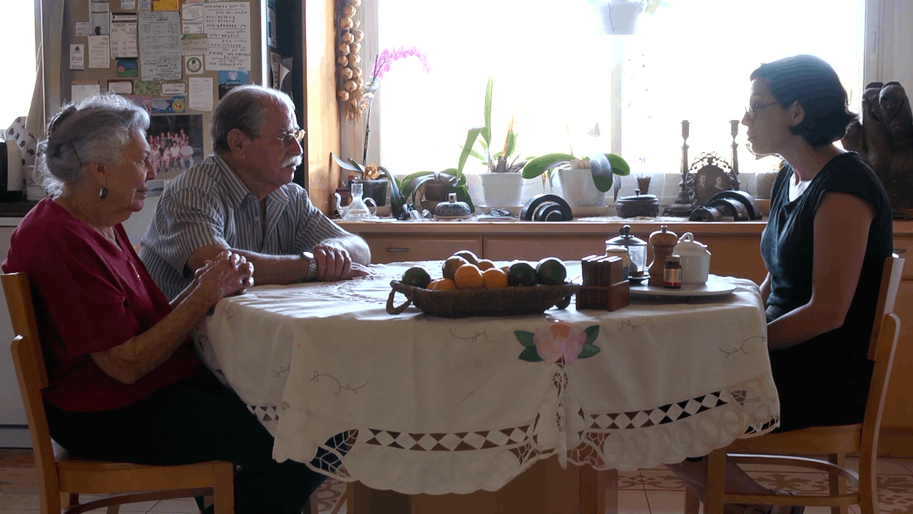 Danielle Schwartz sits at a dining table across from her grandparents in a bright, sunlit kitchen. A bowl of fruit sits between them, and plants and everyday objects line the windowsill behind them.